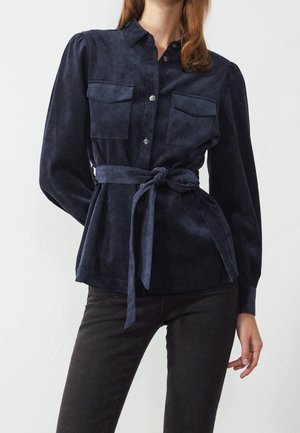Femme portant un chemisier bleu marine boutonné avec des poches à l'avant et une ceinture nouée à la taille, associé à un pantalon noir ajusté.