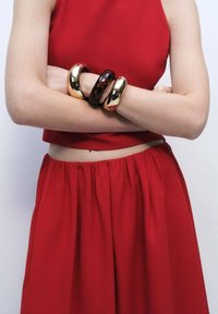 Bracciali in oro e tartaruga sulle braccia incrociate. La persona indossa un top rosso corto e una gonna abbinata con un elastico in vita.