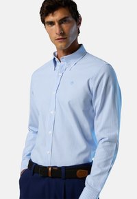 Camisa de botones rayada de color azul claro con cuello puntiagudo. Presenta botones blancos y un logo en el pecho, combinada con un cinturón trenzado negro.