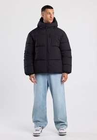 Black puffer jacket with a hood, featuring a zippered front, quilted design, and a chest pocket. Worn over light blue wide-leg jeans.