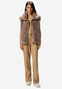 Ovratna jakna iz umetnega krzna v svetlo rjavi barvi z visokim ovratnikom in zadrgo spredaj, nošena preko bež puloverja in bež hlač, kombinirana z belimi supergami.