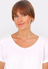 Gul kædetchokker med en lodret stang-vedhæng, der har en glat overflade. Båret over en hvid let T-shirt med scoop-halsudskæring.