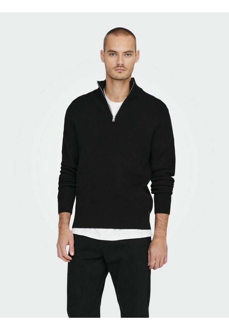 Jersey negro con cuello de cremallera, de manga larga, textura acanalada y corte ajustado. Lleva puesto sobre una camisa blanca, combinado con pantalones negros.