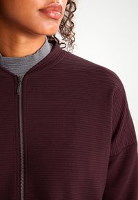 Maroona vetoketjullinen takki, jossa on ribbitekstuuri, korkea kaulus ja metallivetoketju. Kerroksittain harmaan turtleneckin päällä.