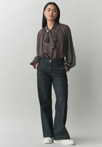 Mustaga polka dotiga blouse, millel on seotav kaelus, kombineeritud kõrge vöökoha tumedad teksapüksid ja valged tennisekingad. Tekstuuriga kangadetailid on nähtavad.