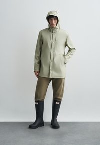 Svijetlo zeleni vodootporni jakna s kapuljačom, prednjim zatvaračem na drukere i bočnim džepovima, uparena s smešnim skraćenim hlačama i crnim gumiranim čizmama.