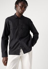 Camisa negra de botones con dos bolsillos en el pecho y un pequeño detalle de logo. Hecha de tela suave, con mangas largas y un cuello clásico.