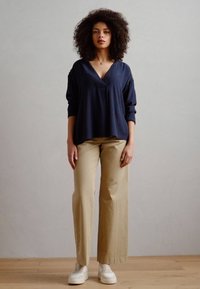 Blusa blu navy a V con maniche lunghe, abbinata a pantaloni larghi beige chiaro e sneakers bianche, su uno sfondo neutro.