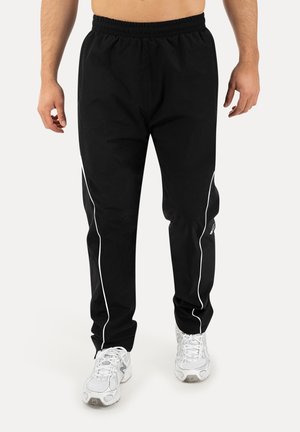 Mężczyzna ubrany w czarne spodnie dresowe z białym paskiem po bokach i białe trampki, stojący na jednolitym tle.