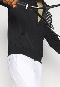 Person i svart jacka och vita byxor som håller och svingar ett svart tennisracket mot en enfärgad bakgrund.