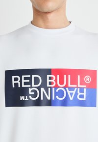 Sudadera blanca con un audaz diseño gráfico multicolor que presenta "RED BULL RACING" en texto azul y rojo sobre un fondo azul marino oscuro.
