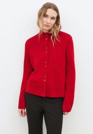 Femme aux longs cheveux blonds portant un cardigan rouge boutonné et un pantalon noir, se tenant devant un fond blanc uni.