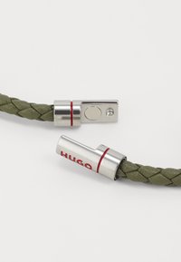 Bracciale intrecciato in pelle verde con chiusura metallica dai dettagli rossi e logo "HUGO" inciso. Texture liscia, design minimalista.