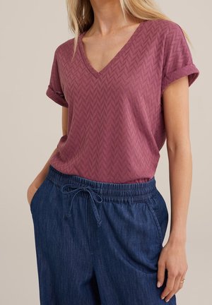 Vrouw draagt een textuurige mauve V-hals top met korte mouwen en omgeslagen manchetten, gecombineerd met blauwe broek met elastische taille en aantrekkoord.