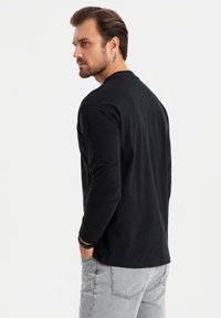 Schwarzes Langarmshirt aus weichem Stoff mit rundem Halsausschnitt. Das Model trägt hellgraue Jeans und zeigt ein Tattoo an seinem Hals.