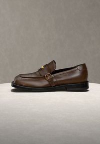 Mocassin en cuir marron avec détail de boucle dorée sur surface beige et fond neutre, vue de côté.