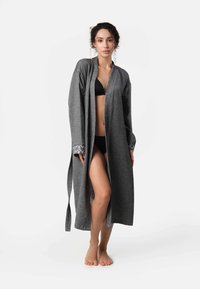 Dlhý šedý župan z jemnej tkaniny s otvoreným predným dielom a širokými rukávmi, s vzorovaným lemovaním na manžetách. Nosený cez čierny bikini.