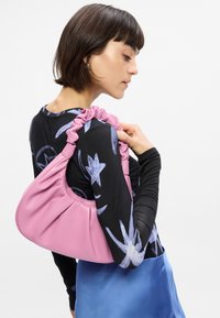 Ροζ, μαλακή δερμάτινη τσάντα με πιασμένο λουρί ώμου, κρατημένη στον ώμο. Το φόντο είναι απλό λευκό, τονίζοντας το σχέδιο της τσάντας.