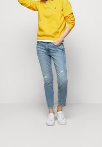 Gul huvtröja med dragsnörningar över en vit skjorta, kombinerad med ljusblå slitna jeans och vita sneakers.