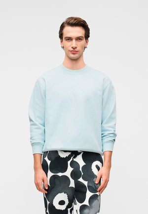 Ανοιχτό μπλε φούτερ με στρογγυλή λαιμόκοψη και ανάγλυφο λογότυπο "marimekko", συνδυασμένο με μαύρο και άσπρο παντελόνι με λουλουδάτο σχέδιο.