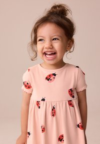 Rosa klänning med korta ärmar, texturerat tyg med mönster av röda och svarta nyckelpigor, veckad kjol och volangkantade ärmslut.