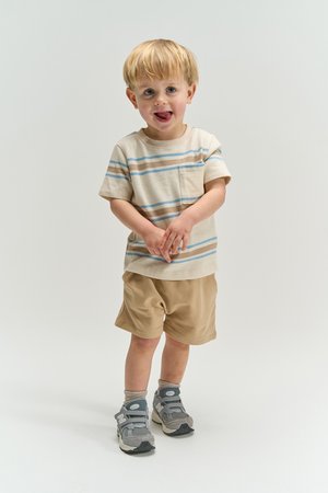 Young boy in beige striped t-shirt and shorts, gray sneakers, standing and smiling with tongue out on a plain white background.