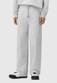 Helle graue Jogginghose aus weichem Material, mit einem Kordelzugbund, weiten Beinen und sichtbaren Nahtdetails. Kombiniert mit weißen und schwarzen Schuhen.