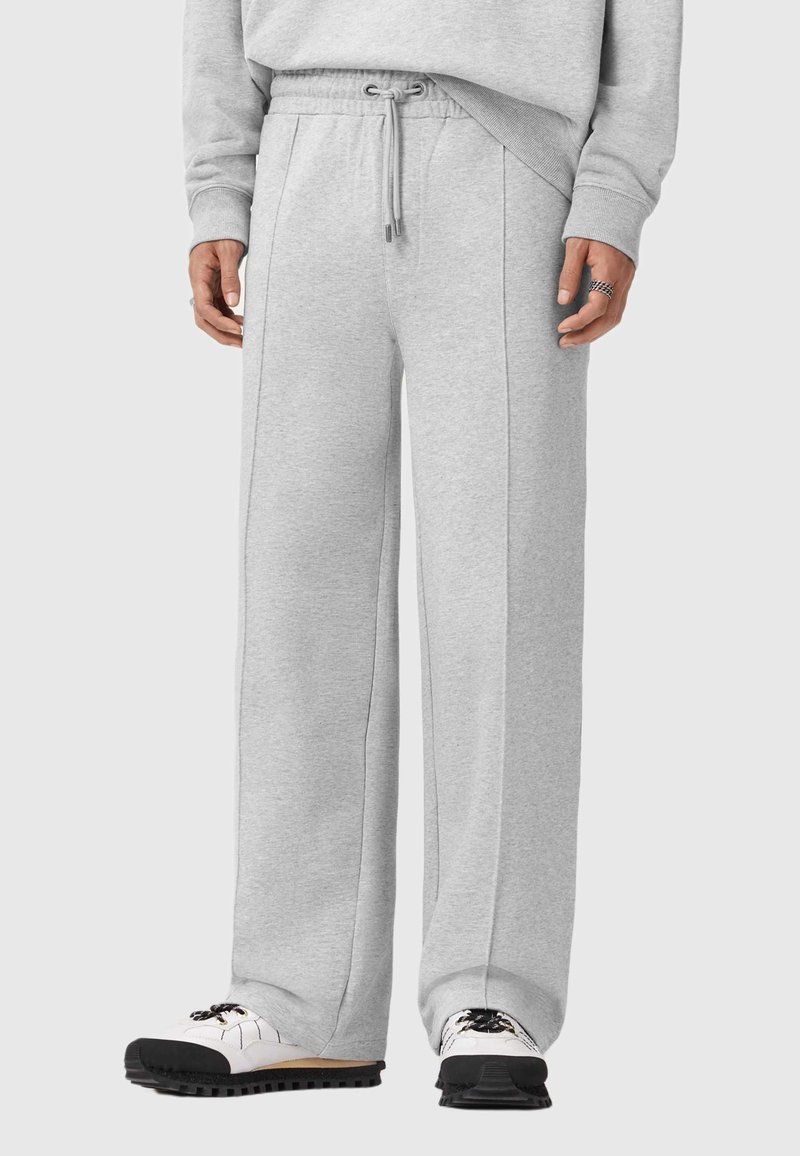 Helle graue Jogginghose aus weichem Material, mit einem Kordelzugbund, weiten Beinen und sichtbaren Nahtdetails. Kombiniert mit weißen und schwarzen Schuhen.