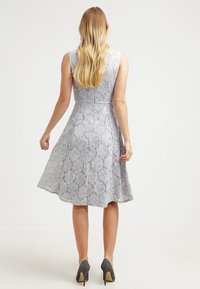 Kvinna med långt blont hår klädd i en ärmlös silverfärgad spettsklänning i A-linjeform och grå högklackade skor, står vänd bortåt mot en vit bakgrund.