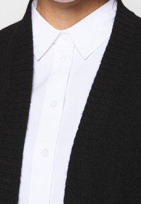 Czarny kardigan z prążkowanej dzianiny nałożony na białą koszulę z kołnierzykiem z guzikami, charakteryzujący się gładką fakturą i kontrastującymi kolorami.