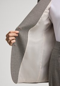 Geruite blazer met een gestructureerd ontwerp, voorzien van een beige binnenvoering, subtiele stikdetails, en een hand die de revers vasthoudt.