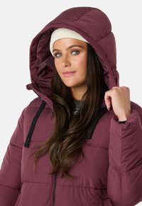 Bordeaux pufferjakke med en stor hætte, sorte snore og lynlås lukning. Båret over en grå sweater med en glat, skinnende tekstur.