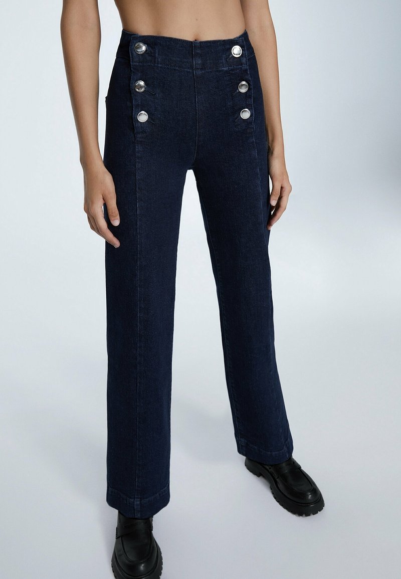 Jean bleu foncé taille haute avec six boutons argentés, coupe droite, porté avec des chaussures noires à semelles épaisses sur fond uni.