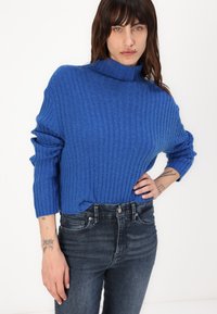 Ribbete blå genser med høy hals og trekvart ermer, sammen med mørke denimjeans. Merkelig tekstur og avslappet passform.