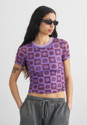 Hosszú sötét hajú nő fekete napszemüvegben, lila mintás crop topot és szürke melegítőnadrágot visel, kezét a zsebében tartva.