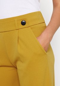 Gelbe maßgeschneiderte Hosen mit strukturiertem Finish, ausgestattet mit einem schwarzen Knopf, Falten und seitlichen Taschen für Funktionalität.