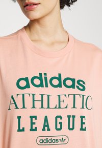 Rožinė medvilninė marškinė su žaliais užrašais, kuriuose rašoma "adidas ATHLETIC LEAGUE", ir logotipu. Rygos apykaklė prideda tekstūros.