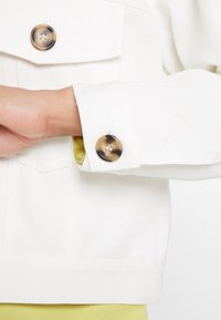 Hvid denimjakke med en knappetærm med to store, glinsende knapper. Stoffet har en let tekstur, og en anelse gult kan ses nedenunder.