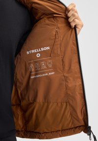 Interno di una giacca a vento marrone con logo bianco del brand e istruzioni per la cura. Tessuto liscio e leggero con una tasca con zip e cordino.