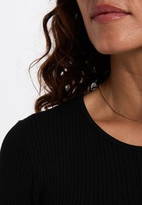 Schwarzes, geripptes Oberteil mit rundem Ausschnitt, ergänzt durch eine zarte goldene Kettenhalskette. Lockige braune Haare teilweise sichtbar.