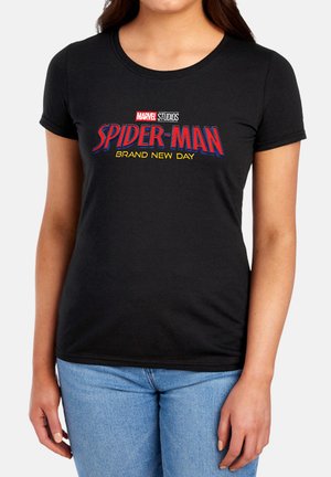 Frau trägt ein schwarzes T-Shirt mit rotem und gelbem Spider-Man Brand New Day-Logo und hellblauen Jeans vor hellem Hintergrund.