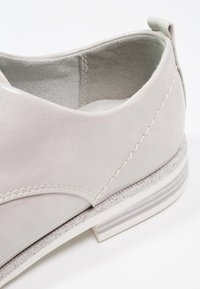 Chaussure gris clair avec un dessus en daim lisse, des coutures décoratives et une semelle texturée. Présente un petit talon avec des accents rayés subtils.
