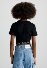 Černé zkrácené tričko s logem Calvin Klein Jeans na pásku, v kombinaci s vysokými modrými džíny z denimu s kapsami na nášivky.
