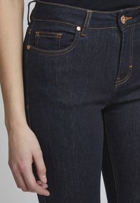 Jeans de denim oscuro con costuras naranjas, que cuentan con un bolsillo frontal, un diseño ajustado y un cierre de botón metálico en la cintura.