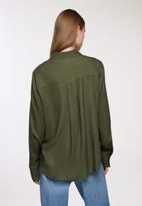 Blusa verde scuro a maniche lunghe realizzata in tessuto morbido, con piega sul retro, colletto e polsini con bottoni. Indossata con jeans azzurri chiari.
