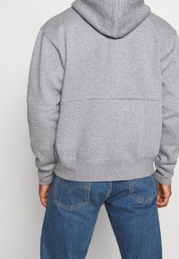 Felpa grigia con tessuto testurizzato, dotata di un ampio cappuccio, spalle scese e una cucitura sul retro. Abbinata a jeans in denim blu.