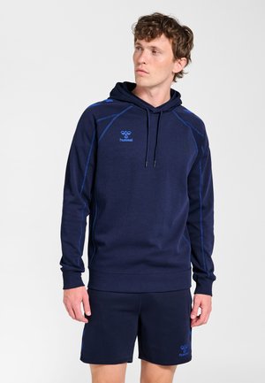 Man in een marineblauwe hoodie met lichtblauwe stikdetails en bijpassende marineblauwe korte broek, staand tegen een effen lichte achtergrond.