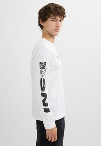 Camiseta blanca de manga larga con texto gráfico negro en las mangas. Hecha de una tela ligera, presenta un cuello redondo y un corte slim.