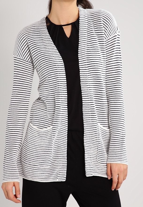 Femme portant un cardigan blanc à fines rayures noires horizontales par-dessus un haut noir, les mains détendues le long du corps.