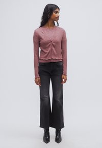 Πλεκτό ροζ τοπ με μακριά μανίκια και λεπτομέρεια με κόμπο μπροστά, συνδυασμένο με ψηλόμεσα μαύρα τζιν με καμπάνα και μαύρες μυτερές μποτάκια αστραγάλου.